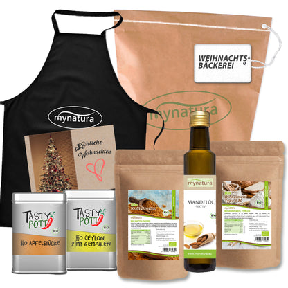 Set aus verschiedenen Bio-Produkten zum Backen und Kochen, darunter Mehle, Mandelöl, Dattelzucker, Apfelstücke, Zimt, eine schwarze Schürze und eine braune Papiertüte mit Weihnachtsmotiv.