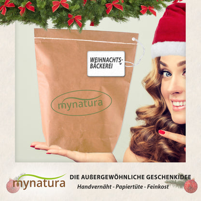 Handvernähte Papiertüte mit Mynatura Logo und Anhänger Weihnachtsbäckerei, von einer Frau mit Weihnachtsmütze präsentiert, als Geschenkidee für Feinkost Sets.