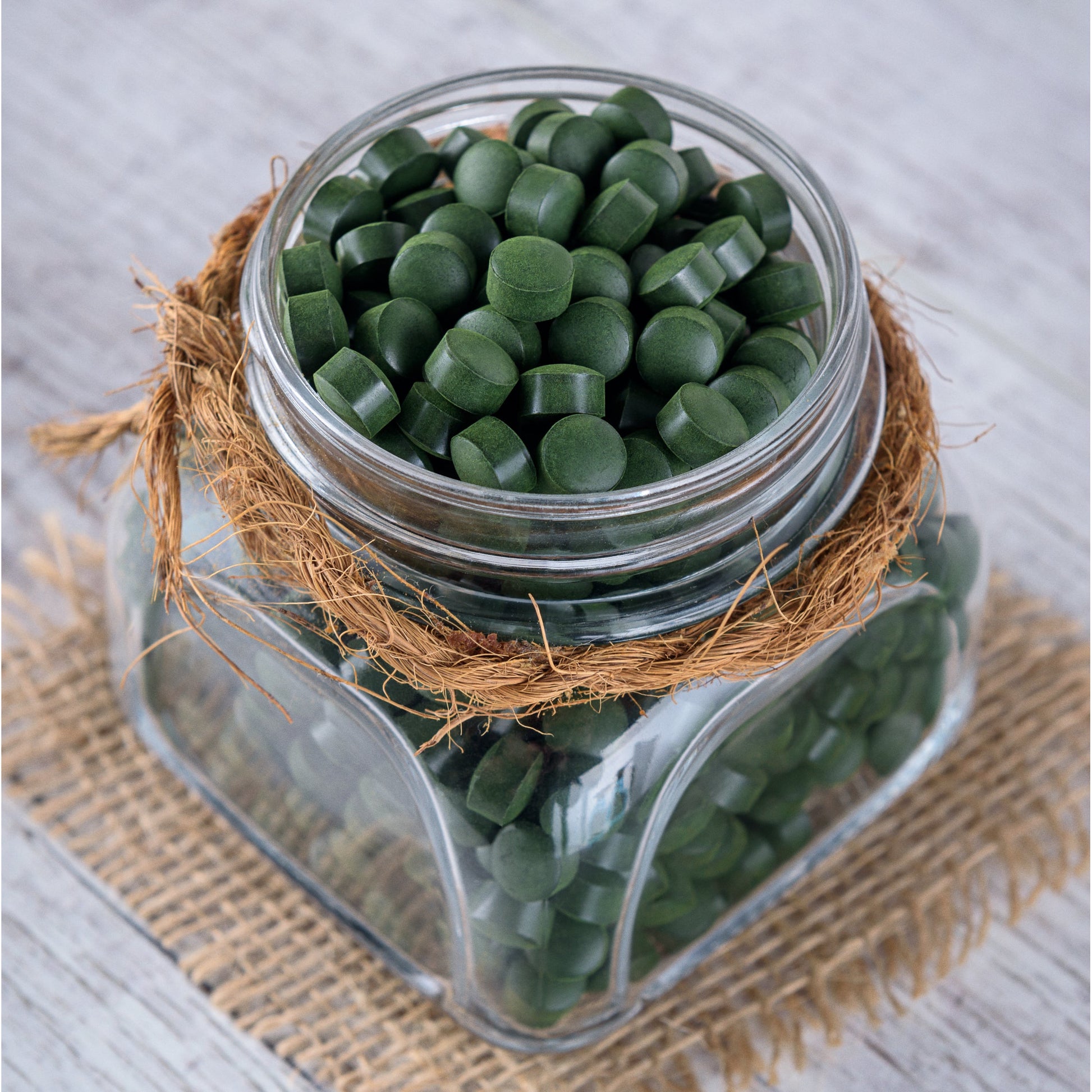 Glasbehälter mit vielen grünen spirulina chlorella algen tabletten, dekoriert mit Bast, auf einem Jutestoff stehend.
