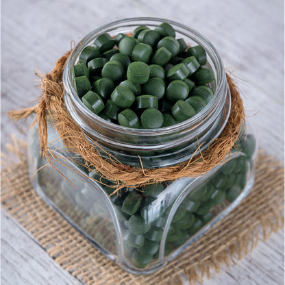 Glasbehälter mit vielen grünen spirulina chlorella algen tabletten, dekoriert mit Bast, auf einem Jutestoff stehend.
