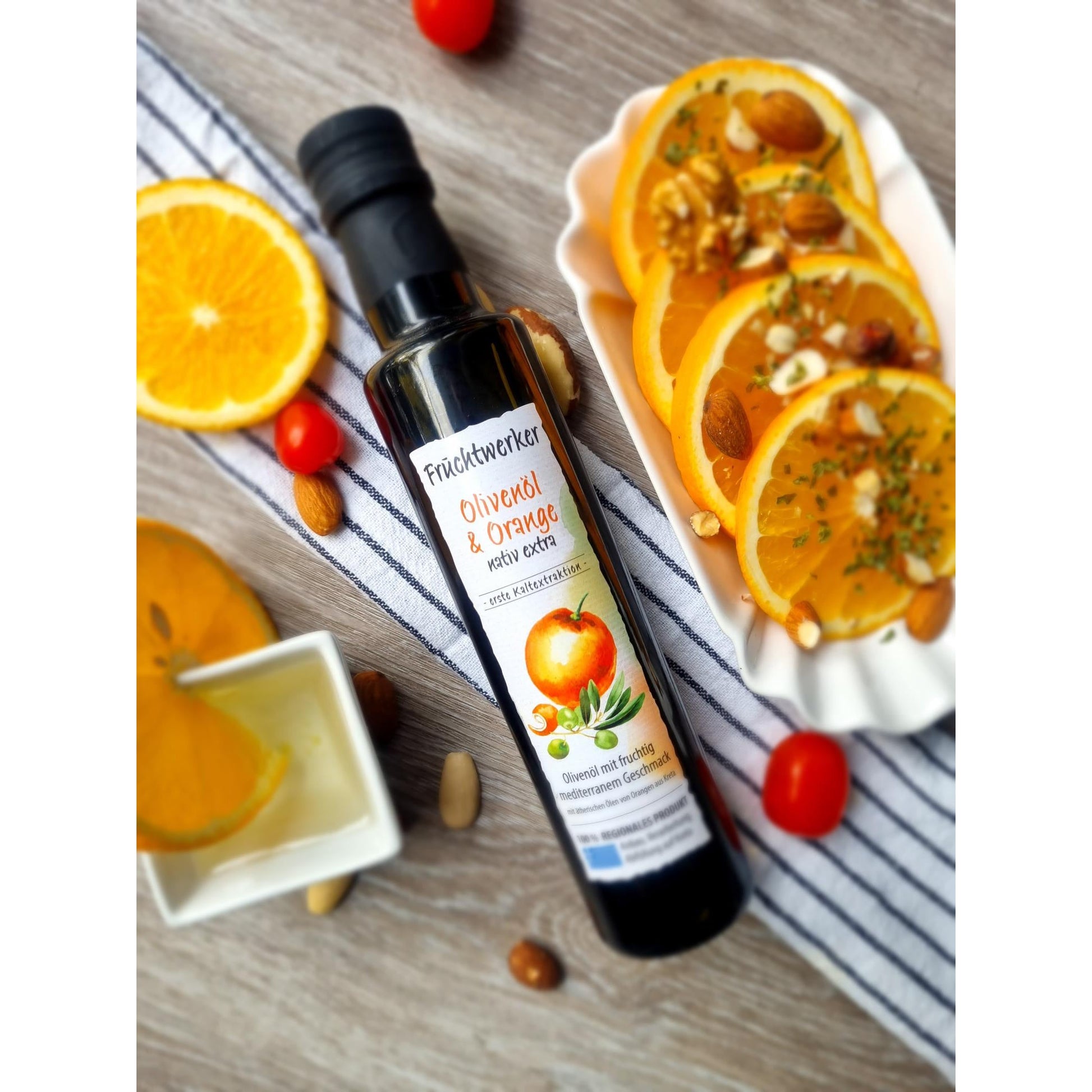 Olivenöl + Orange 250ml von Fruchtwerker auf gestreiftem Tuch, umgeben von Orangenscheiben, Nüssen, Tomaten und einem Schälchen Öl, Szene für Essig & Öle.