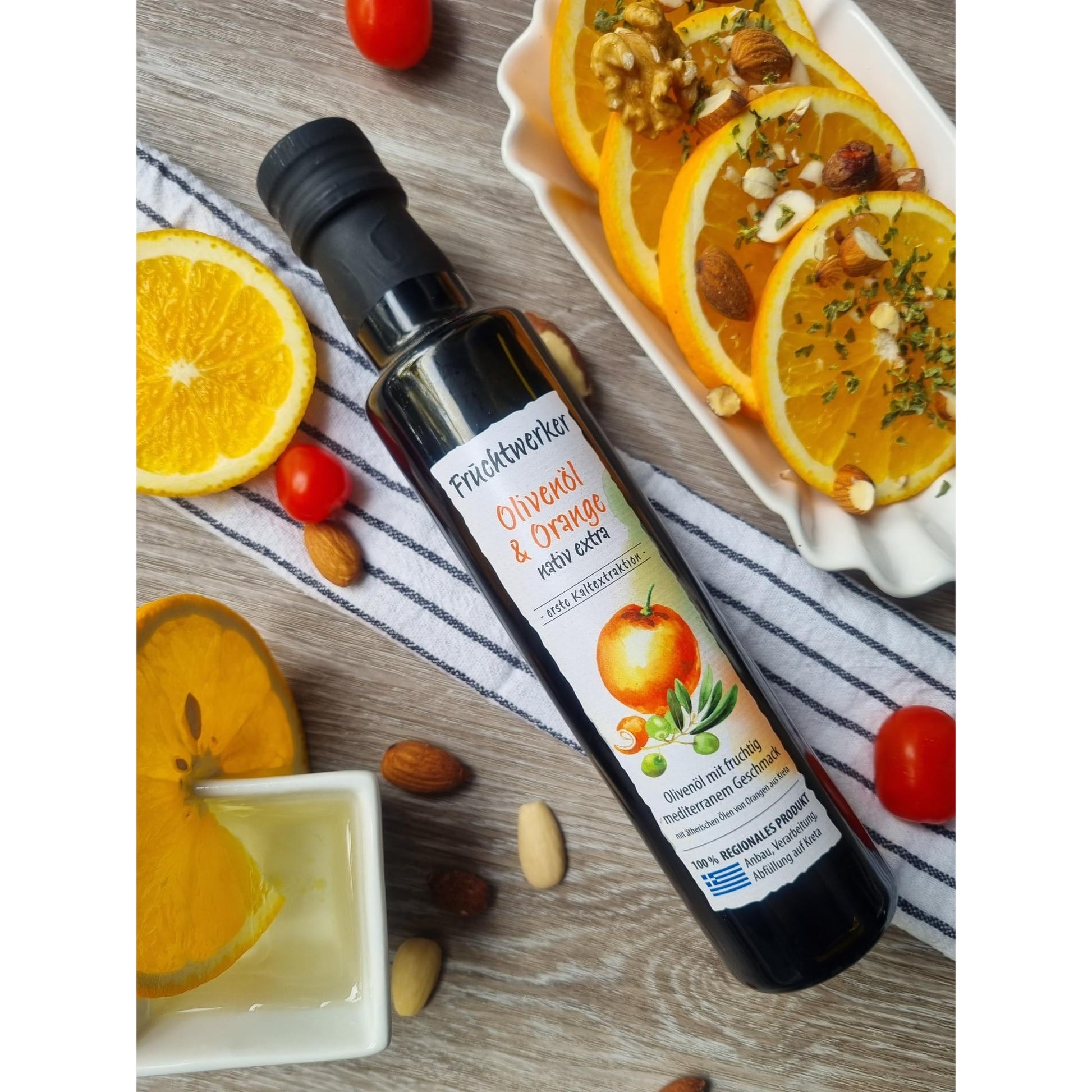 Fruchtwerker Olivenöl + Orange 250ml auf Holz, neben Orangenscheiben mit Nüssen, Tomaten, Mandeln und einem Schälchen Öl, Arrangement für Öle & Säfte.