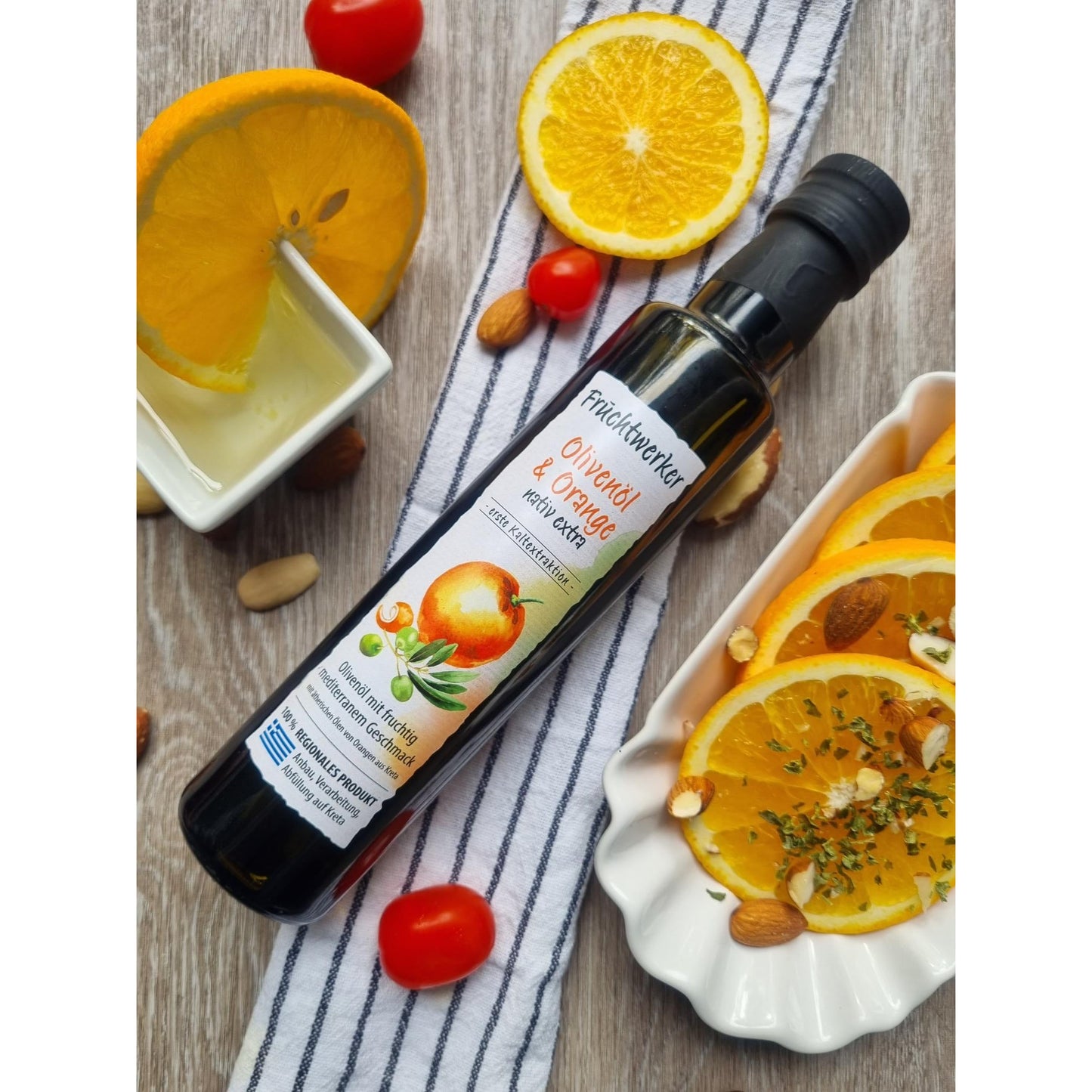 Flasche Fruchtwerker Olivenöl + Orange 250ml auf gestreiftem Tuch, umgeben von Orangenscheiben, Tomaten, Nüssen und einem Schälchen Öl, geeignet für Kochen.