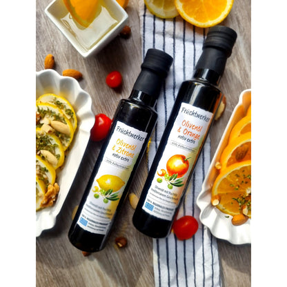 Zwei Flaschen Fruchtwerker Olivenöl, eine mit Zitrone und eine mit Orange, 250ml, auf einem Holztisch zwischen frischen Zitronen- und Orangenscheiben, Nüssen und Tomaten arrangiert.