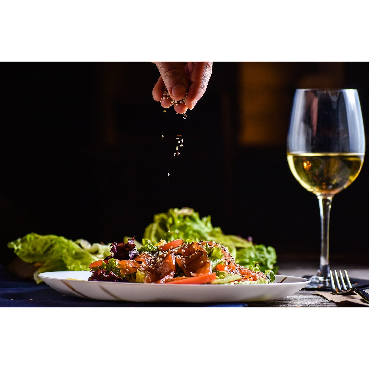 Salatteller mit Gemüse und Topping, darüber werden Samen gestreut, im Hintergrund ein Glas Weißwein