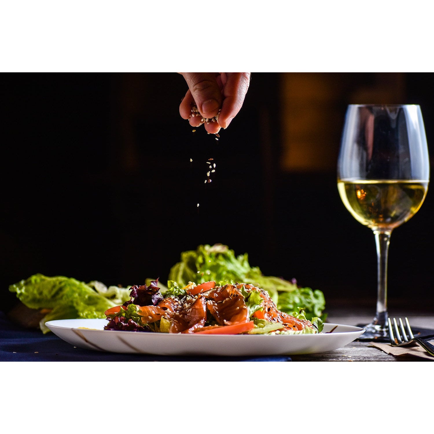 Salatteller mit Gemüse und Topping, darüber werden Samen gestreut, im Hintergrund ein Glas Weißwein