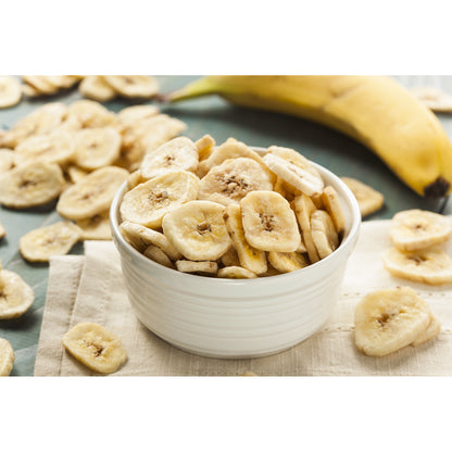 Schale mit knusprigen Bananenchips als Trockenfrucht, im Vordergrund auf einem Tuch, im Hintergrund eine frische Banane und weitere Chips verstreut.
