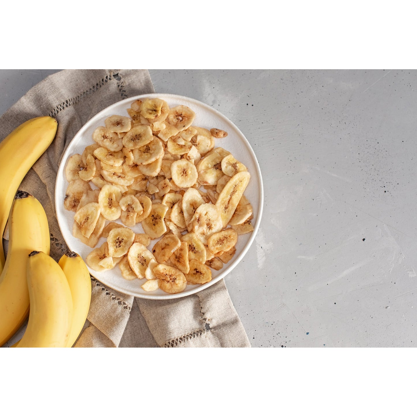 Teller mit Bananenchips als Snacks, daneben mehrere frische Bananen und ein beiges Tuch auf grauem Untergrund.