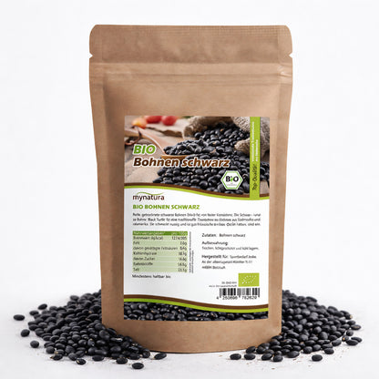 Beutel mit Bio schwarze Bohnen von mynatura, stehend auf einer Unterlage aus verstreuten schwarzen Bohnen, Vorderseite mit Etikett sichtbar.