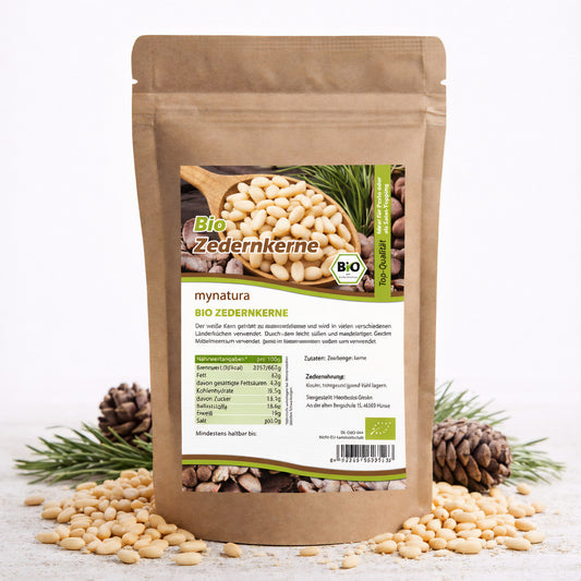 Beutel mit Bio Zedernkerne von Mynatura, 500g, stehend auf weißem Untergrund, umgeben von Zedernnüsse, Kiefernzapfen und grünen Zweigen.