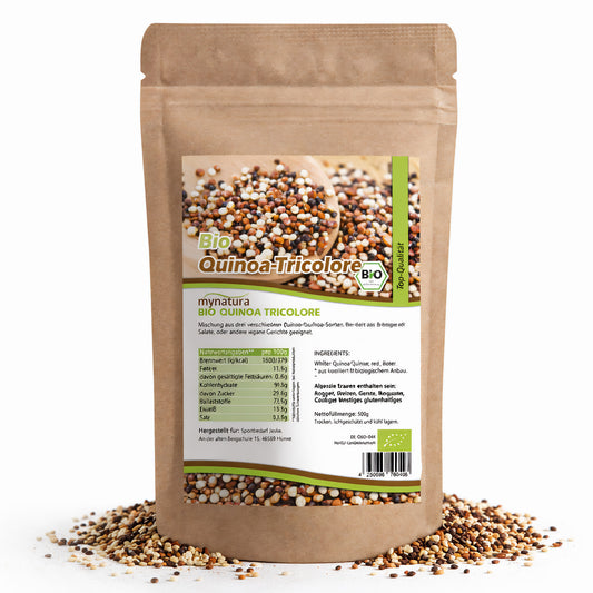 Beutel mit Bio Quinoa Tricolore Mischung, 500g, aus weißen, roten und schwarzen Quinoa-Körnern, stehend auf verstreuten Kernen und Samen.