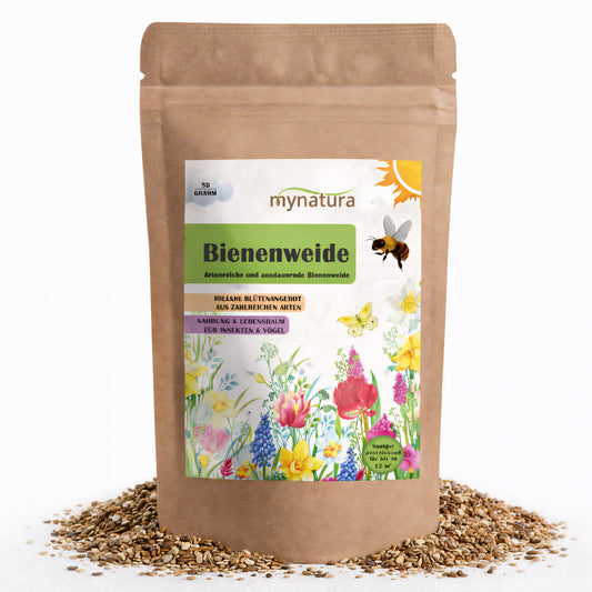 Beutel Mynatura Bienenweide bunte Mischung 50g auf weißem Hintergrund, umgeben von verstreuten Samen, Vorderseite mit Blumenillustration und Produktlabel.