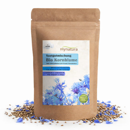 Braune Tüte mit Etikett der Mynatura Bio Kornblume Saatgutmischung, umgeben von verstreuten Kornblumen-Blütenblättern und Blumensamen auf weißem Hintergrund.