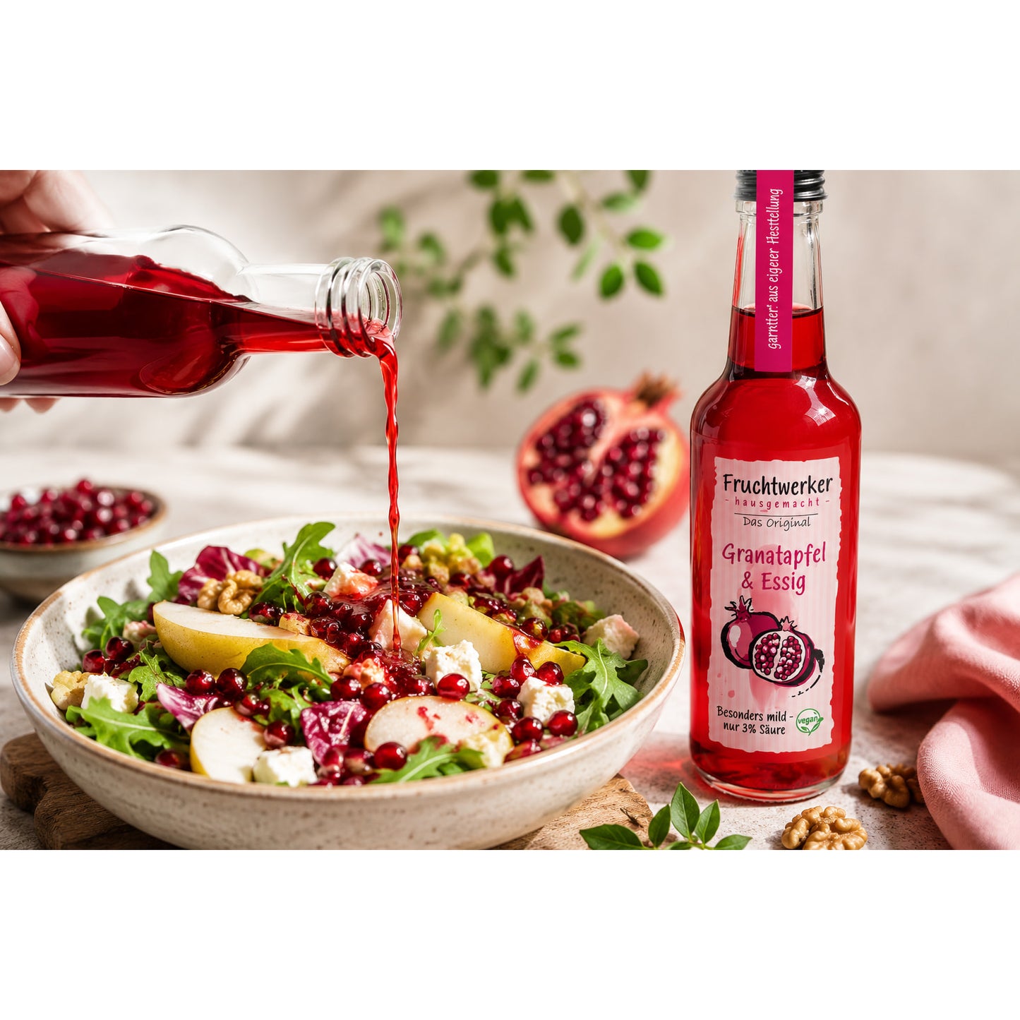 Fruchtwerker Granatapfel & Essig neben einer Schale Salat, aus einer Flasche wird Essig über den Salat gegossen, im Hintergrund Granatapfelhälften.