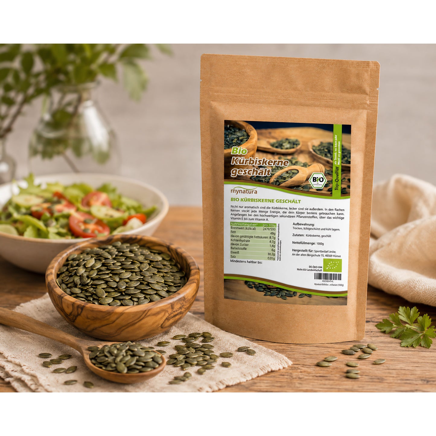 Beutel Bio Kürbiskerne geschält und ungeröstet auf Holztisch, daneben eine Schale mit Kürbiskernen und ein Löffel, im Hintergrund ein Salat.