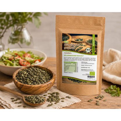 Beutel Bio Kürbiskerne geschält und ungeröstet auf Holztisch, daneben eine Schale mit Kürbiskernen und ein Löffel, im Hintergrund ein Salat.