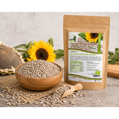 Bio Sonnenblumenkerne, geschält, im Beutel neben einer Holzschale und einem Holzlöffel mit Kernen, dekoriert mit Sonnenblume und Sonnenblumenkopf, auf rustikalem Untergrund.