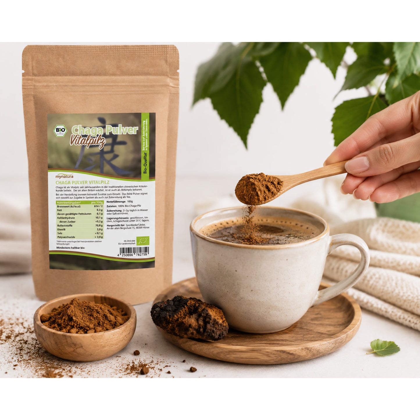 Chagapulver wird mit einem Holzlöffel in eine Tasse mit Getränk gegeben, daneben Beutel Bio Chaga Pulver, Schale mit Pulver und Chaga Pilz auf Holzuntersetzer.