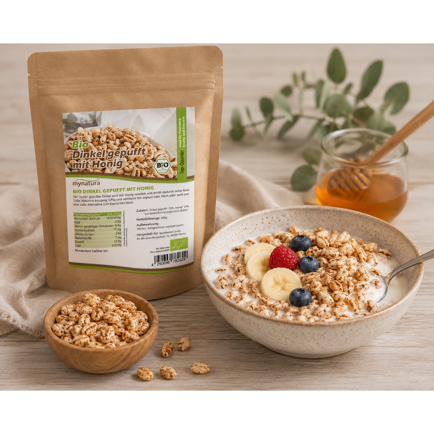 Bio Dinkel gepufft mit Honig als Zutat in einer Müslischale mit Joghurt, Banane und Beeren, daneben Beutel und kleine Holzschale.