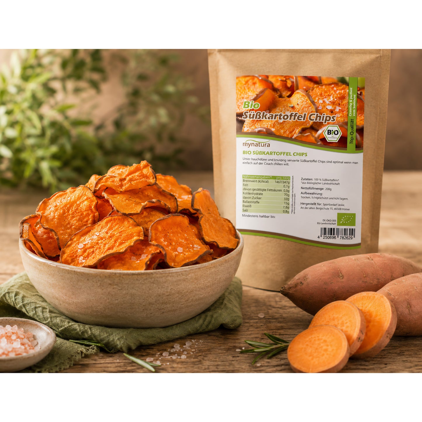 Schale mit knusprigen süßkartoffelchips als snack, daneben Beutel Bio süßkartoffelchips, frische süßkartoffel und Kräuter im Hintergrund