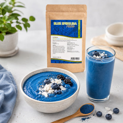 Blaues Spirulina Pulver im Beutel, daneben eine blaue Smoothie-Bowl und ein Glas mit Smoothie, dekoriert mit Beeren und Kokos, auf einem Tisch.