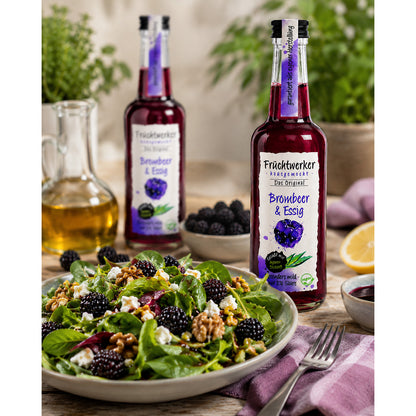 Flasche Fruchtwerker Brombeer & Essig auf Tisch neben Salat mit Brombeeren, Walnüssen und Feta, im Hintergrund weitere Flasche und Zutaten.