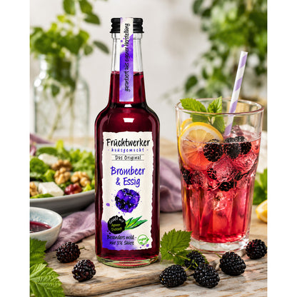 Flasche Fruchtwerker Brombeer & Essig neben Glas mit Brombeer-Getränk, Eiswürfeln, Zitronenscheibe und frischen Brombeeren auf Holztisch.
