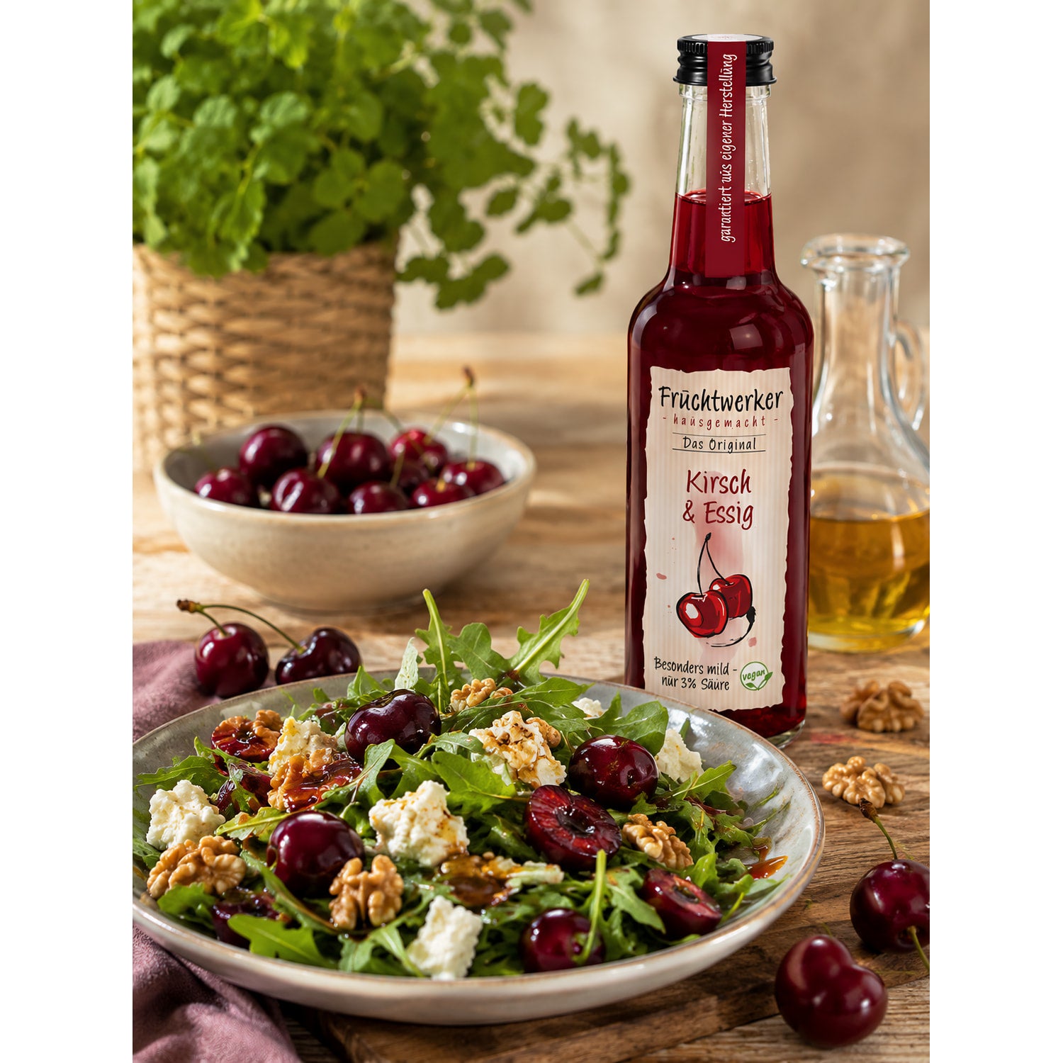 Fruchtwerker Kirsch & Essig 250ml neben einem frischen Salat mit Kirschen, Walnüssen und Feta, im Hintergrund eine Schale Kirschen und Kräuter.