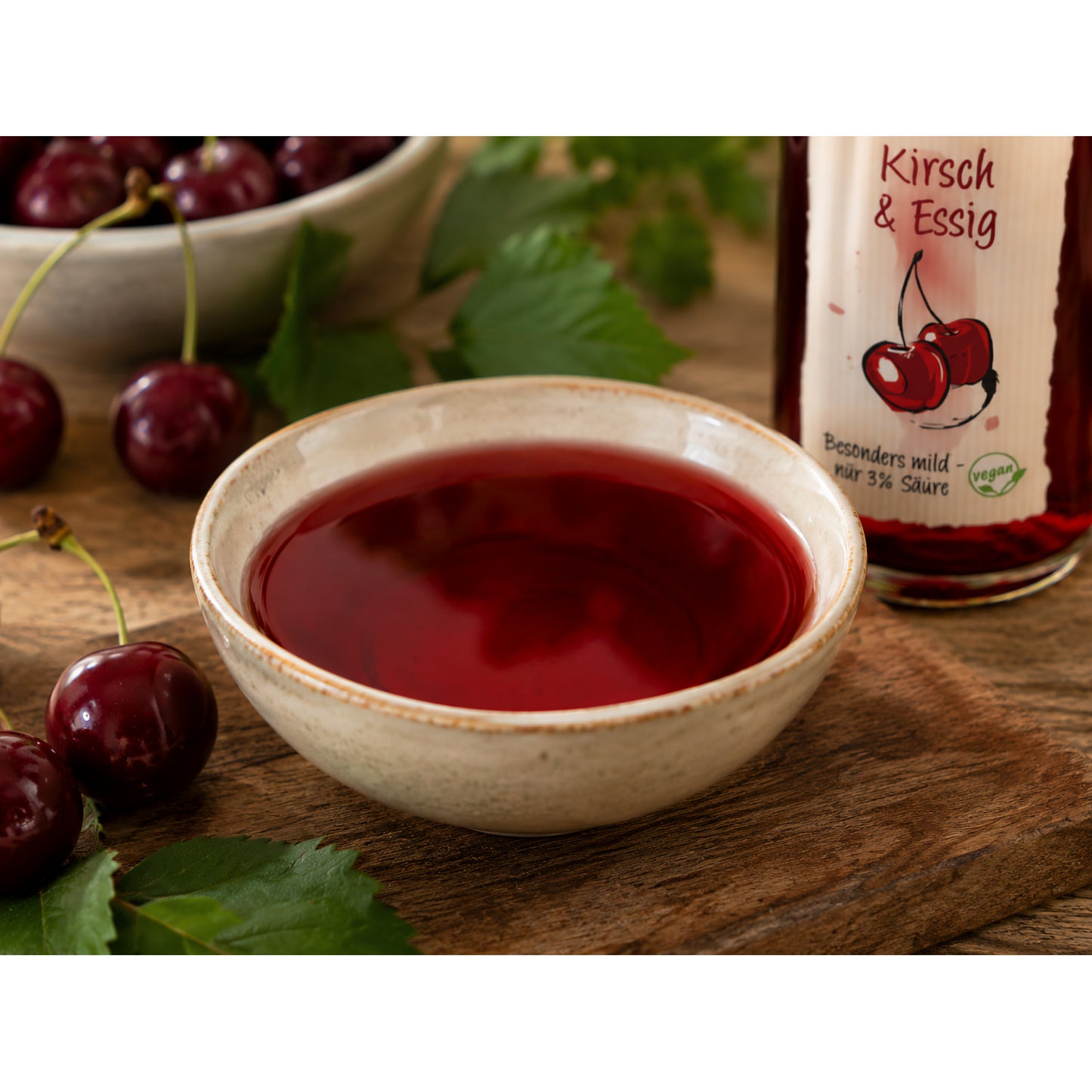 Schale mit rotem Kirsch & Essig, daneben frische Kirschen und ein Teil der Flasche Fruchtwerker Kirsch & Essig 250ml auf Holzuntergrund.