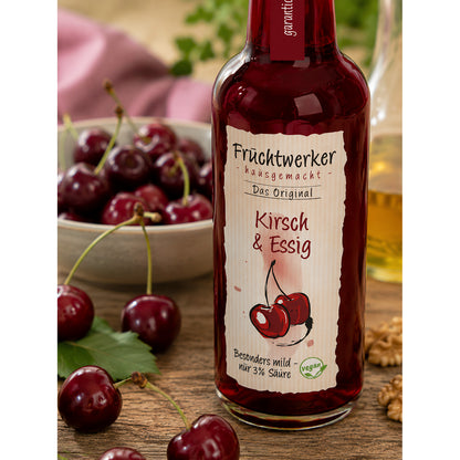 Fruchtwerker Kirsch & Essig 250ml mit Fokus auf das Etikett, im Hintergrund Schale mit Kirschen, Walnüsse und grüne Blätter.