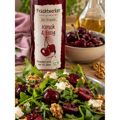 Nahaufnahme einer Flasche Fruchtwerker Kirsch & Essig 250ml vor einem angerichteten Salat mit Kirschen, Walnüssen und Feta.