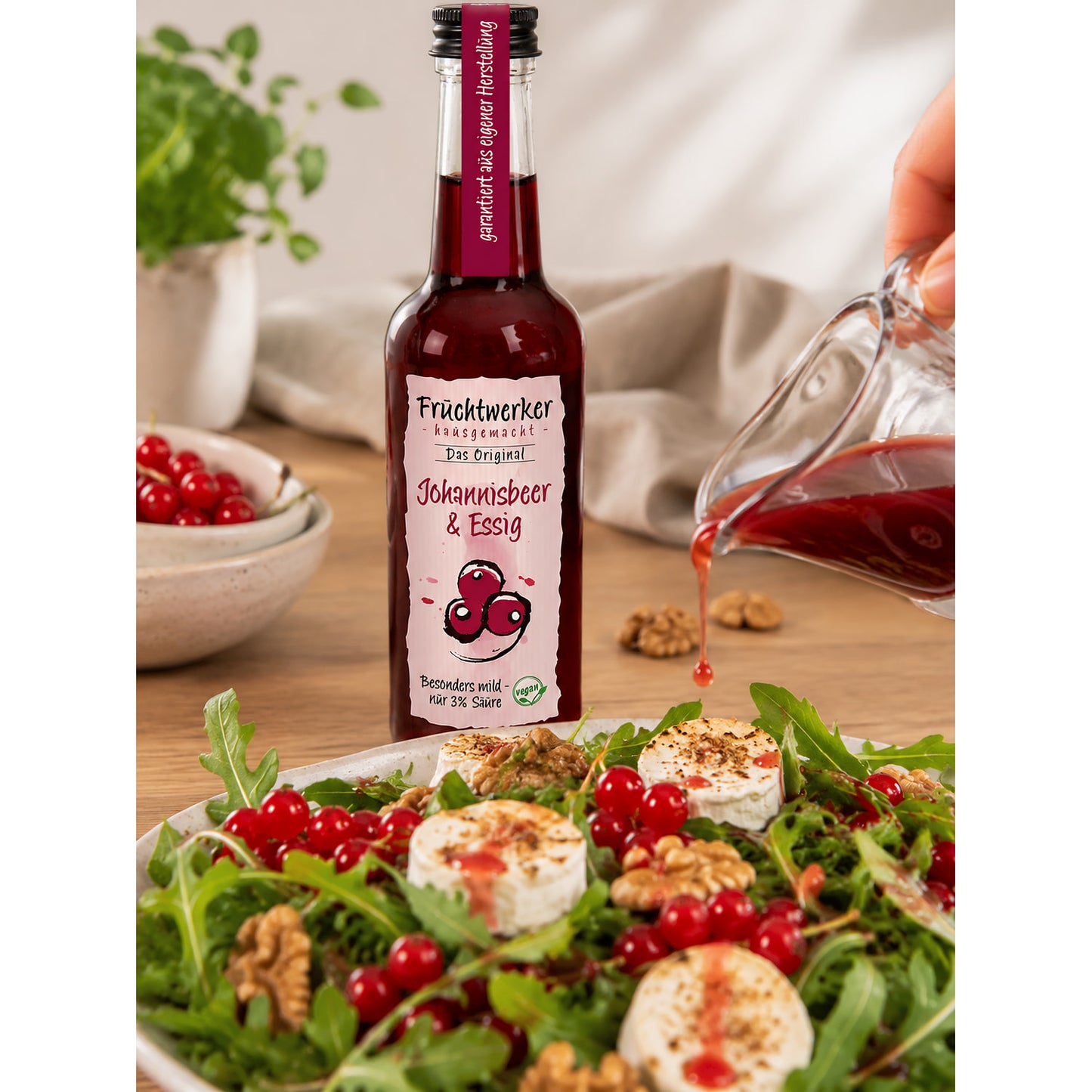 Fruchtwerker Johannisbeer-Essig 250ml Flasche neben Salat mit Walnüssen, Johannisbeeren und Ziegenkäse, Essig wird aus Glaskännchen über den Salat gegossen.