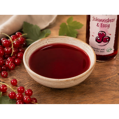 Rote Flüssigkeit aus Fruchtwerker Johannisbeer-Essig in einer Keramikschale, daneben Johannisbeeren und Flaschenausschnitt auf Holzuntergrund.