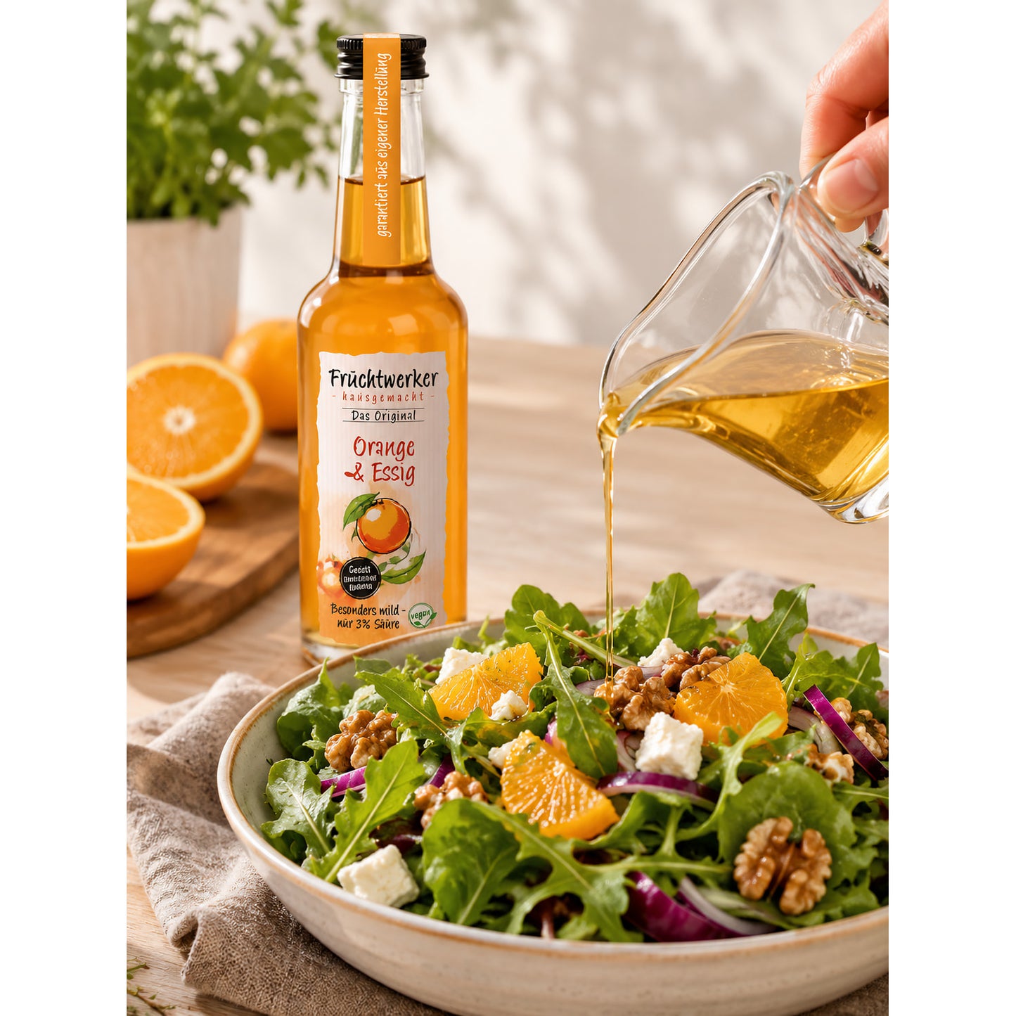 Fruchtwerker Orange & Essig Flasche neben einem Salat, über den aus einer Glaskanne Essig gegossen wird, im Hintergrund Orangen.