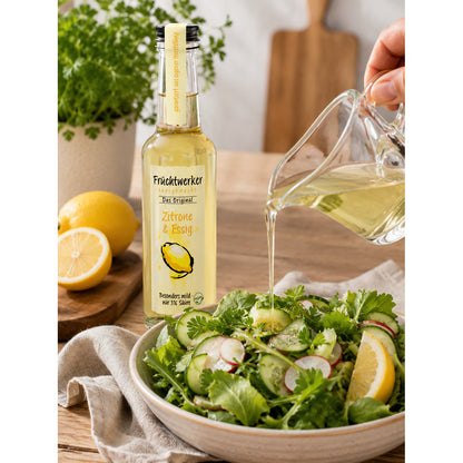 Fruchtwerker Zitrone & Essig Flasche neben Zitronen, während Essig aus einer Glaskanne über einen frischen Salat gegossen wird.