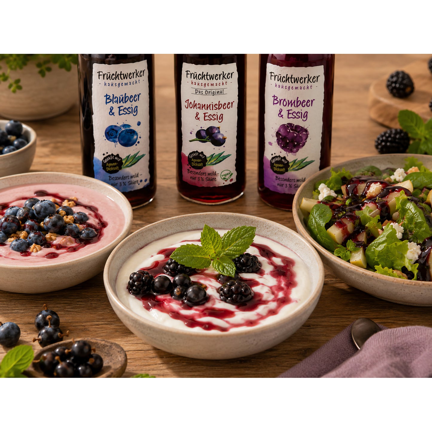Fruchtwerker Essig und Frucht Set mit drei Flaschen im Hintergrund, davor Schalen mit Joghurt, Beeren und Salat, alles auf einem Holztisch angerichtet.
