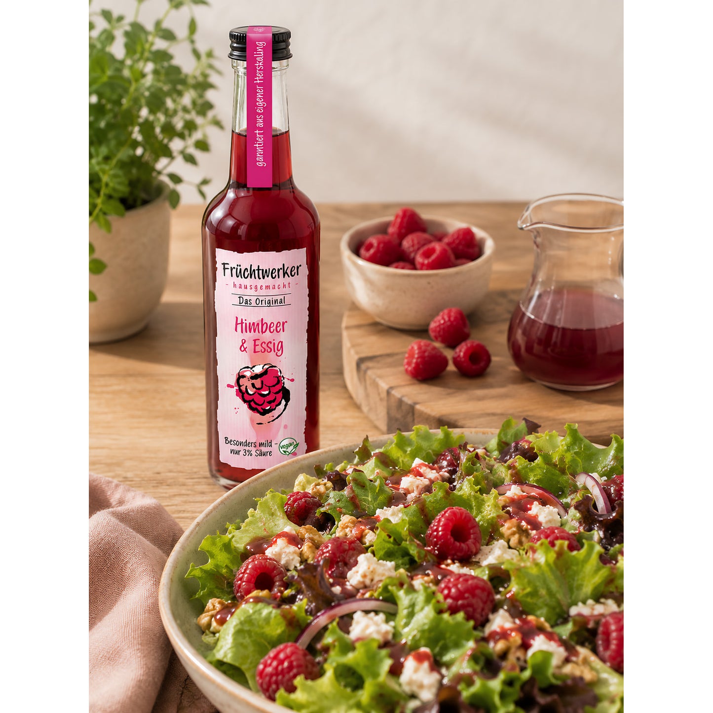 Fruchtwerker Himbeer & Essig 250ml auf einem Holztisch neben frischen Himbeeren, einer Karaffe und einem angerichteten Salat.