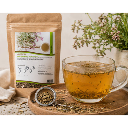 Zubereitungsszene mit einer Tasse Tee aus erdrauchkraut, daneben ein Teesieb mit geschnittenem kraut und der geöffnete Beutel im Hintergrund.